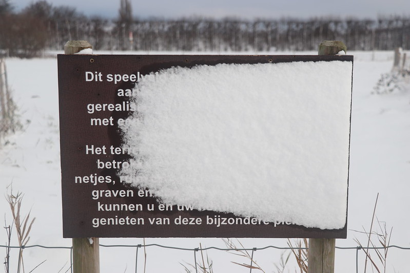 Bordje over speelveld voor honden, alleen 'genieten van deze bijzondere plek' is goed te lezen, voor de rest zit er sneeuw over.